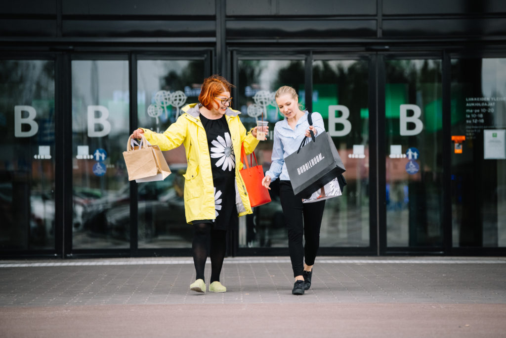 Kaksi naista kantavat ostoskasseja ja astuvat ulos kauppakeskuksesta.