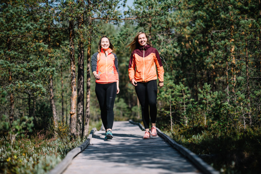 Kaksi metsässä pitkospuilla lenkkeilevää naista katsovat toisiaa. nauraen