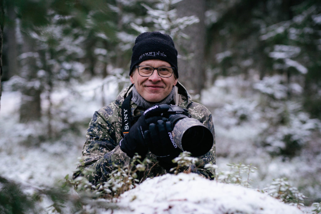 Mies hymyilee iso kamera käsissään. Taustalla luminen metsä.
