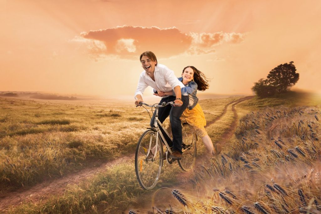 Nuori mies vie naista polkupyörän kyydissä peltotiellä.