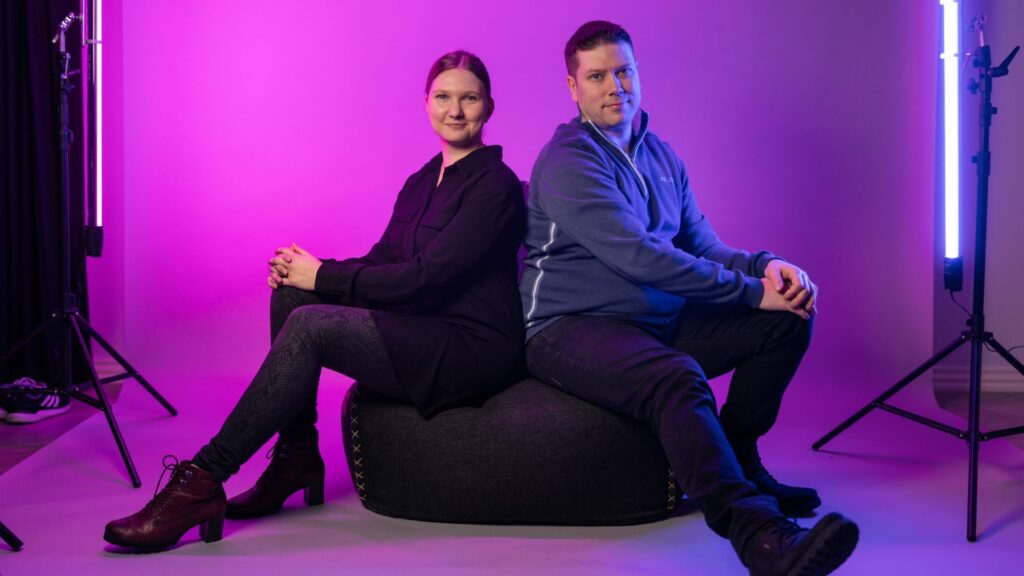 Nainen ja mies istuvat mustalla kuvausstudion tuolilla. Taustalla violetti valo.