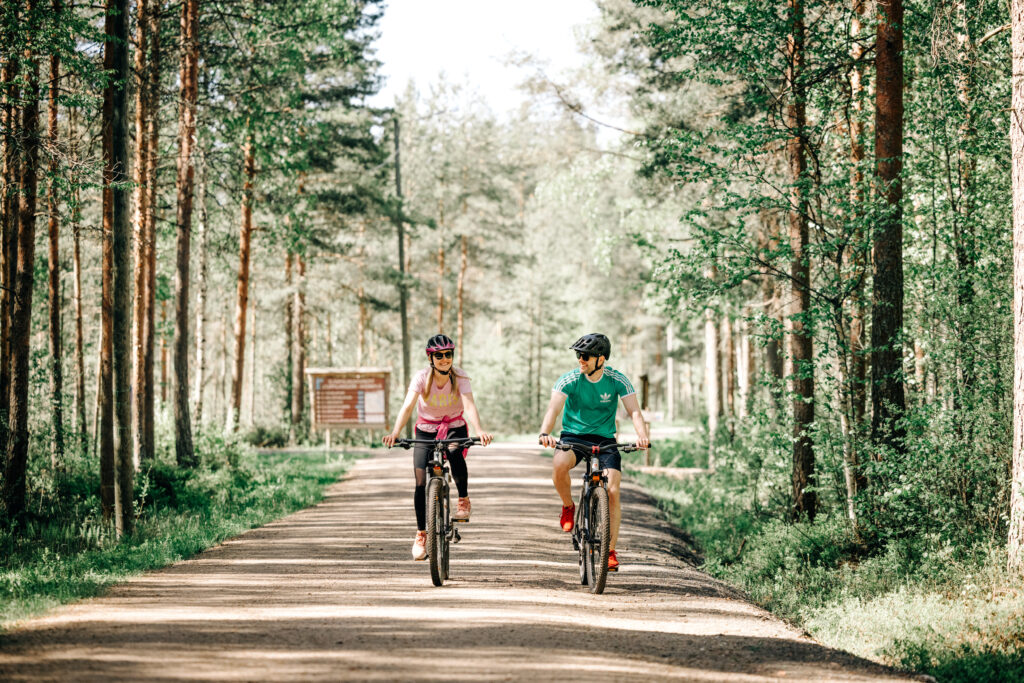 Kaksi henkilöä pyöräilemässä reitillä, jonka ympärillä on metsää.