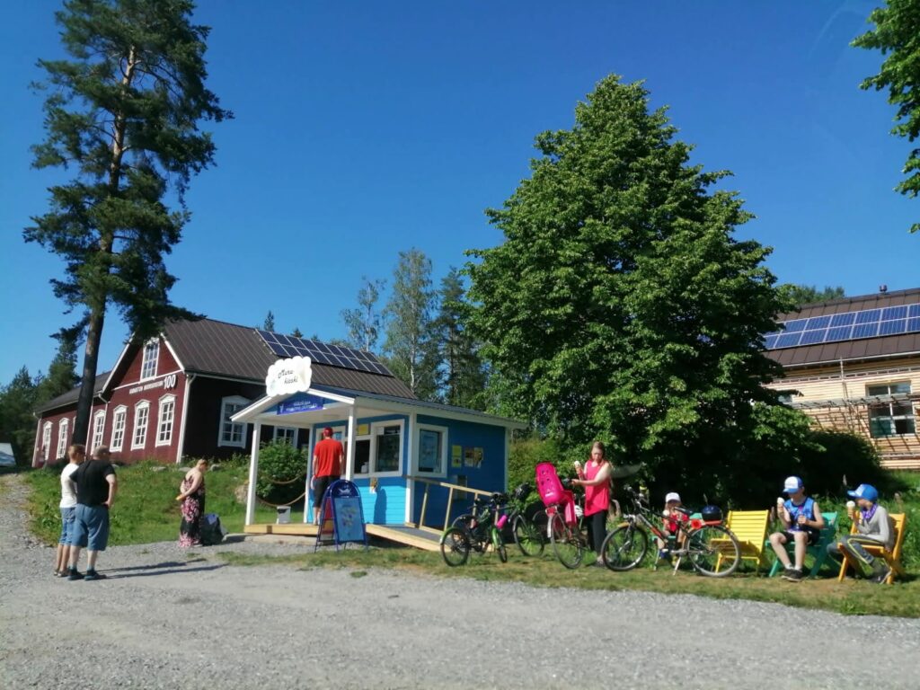 Jäätelökioskin edustalla on ihmisiä ja aurinko paistaa pilvettömältä taivaalta. Taustalla näkyy iso punainen kylätalo. 