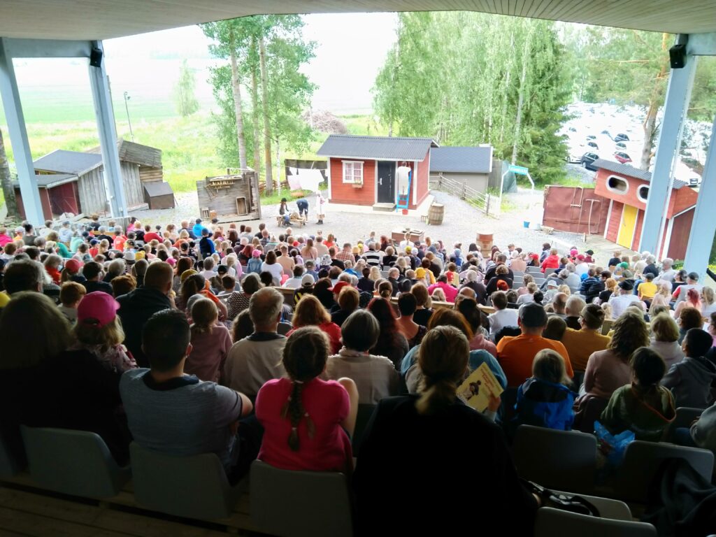 Kesäteatterin katsomossa istuu paljon ihmisiä. Ihmisten edessä lavastettu pihapiiri.