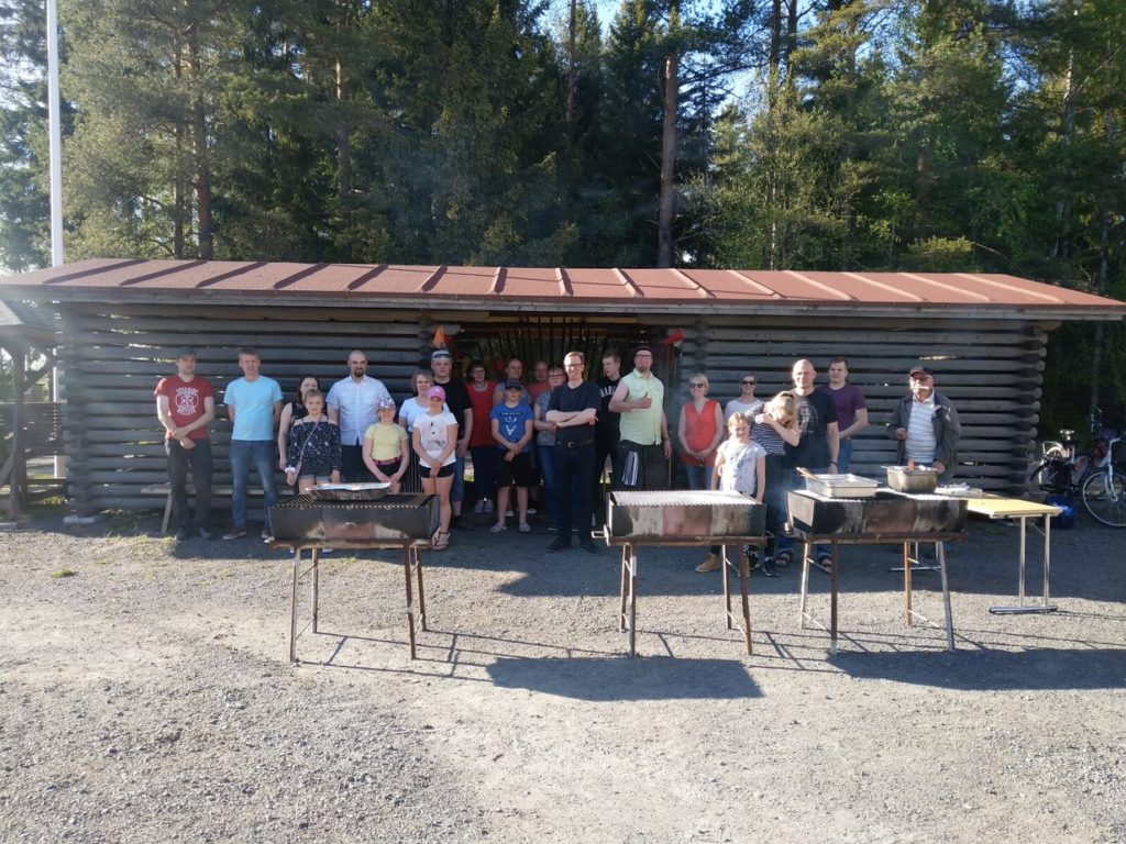 Yli 20 ihmistä seisoo hirsisen rakennuksen edessä. Ryhmän edessä isoja pihagrillejä.