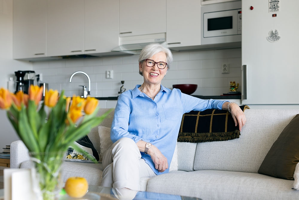 A woman is sitting on a sofa. The kitchen can be seen in the background.