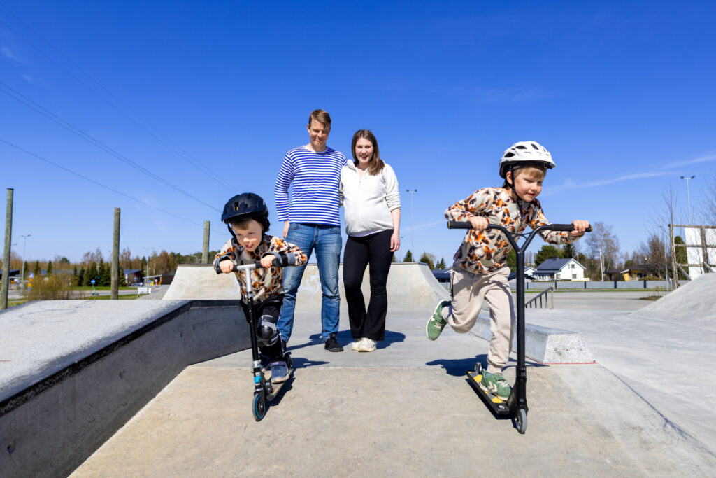 Two young boys in action at a scooter park. Their father and mother stand in the background.