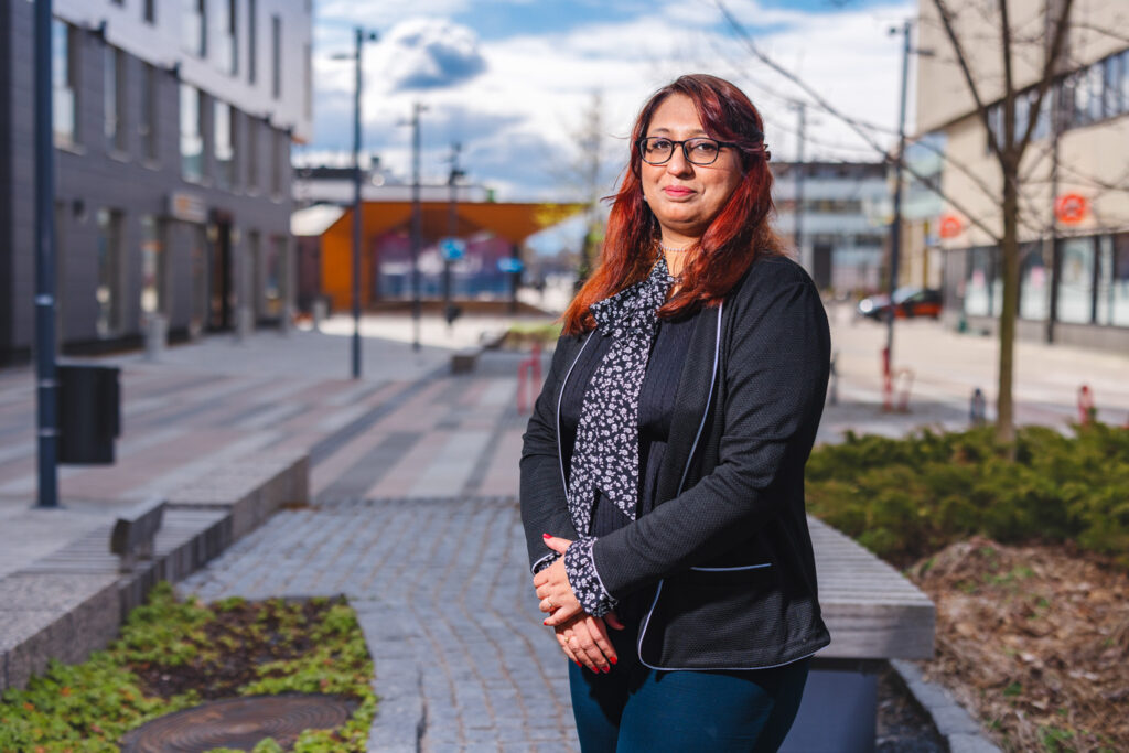 A red-haired woman stands with a park, paving, and apartment buildings visible in the background.