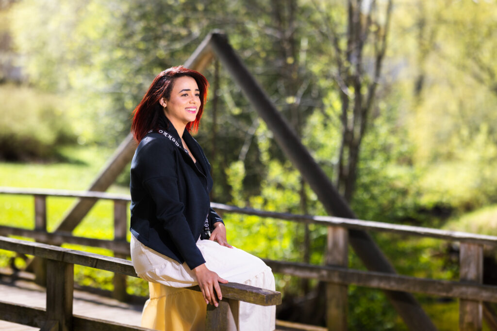 A woman sits smiling on a wooden bridge railing. Green nature and trees are visible in the background.