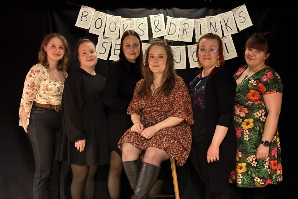 Kuusi naista poseeraavat mustalla taustalla. Naisten takana näkyy teksti: books & drinks Seinäjoki.