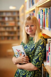 Mervi Heikkilä aikuisten kirjasalin hyllyillä kirja kädessään.