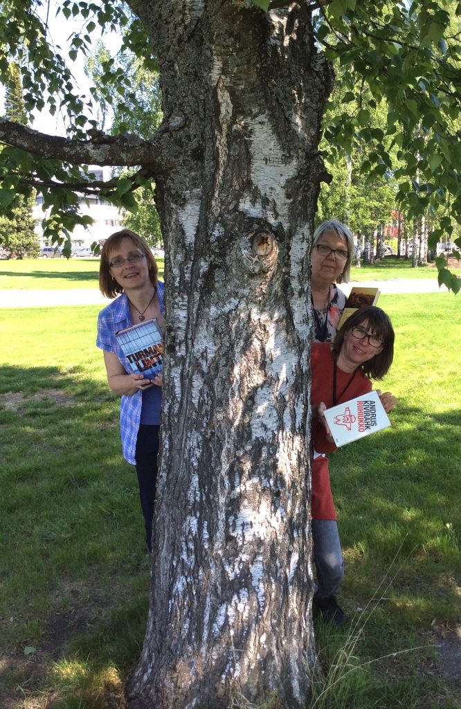 Tiina, Anja ja Anna kurkkivat puun takaa kirjaston pihalla. Jokaisella on kirja kädessään.