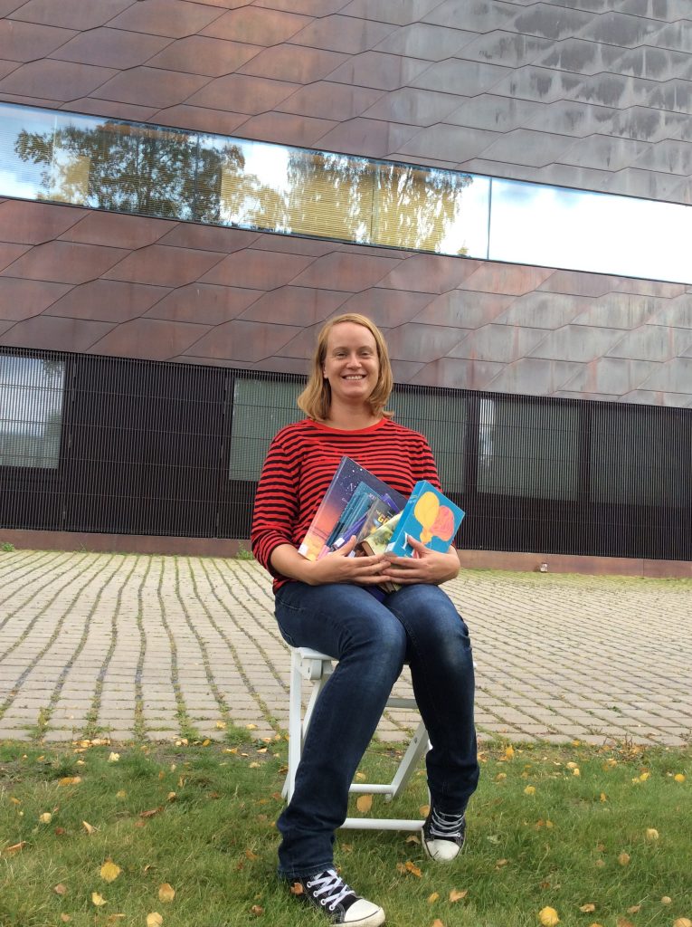 Maria Lehtola istuu Apila-kirjaston edessä pihalla kirjakasa sylissä.