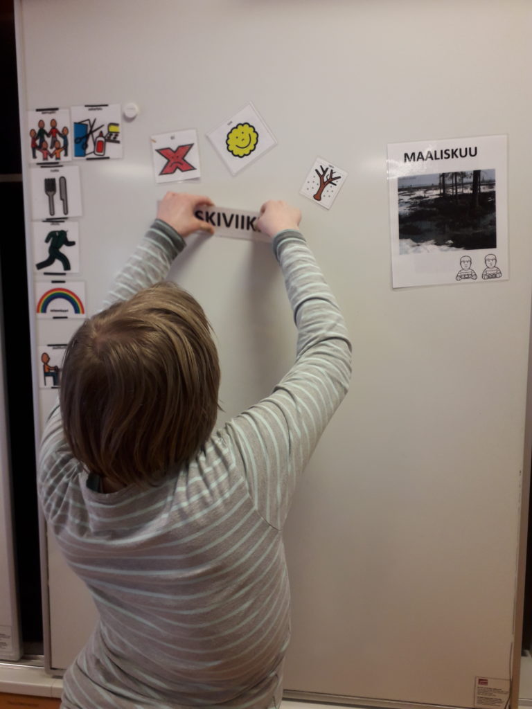 Lapsi kiinnittämässä viikonpäivä-lappua magneetilla seinään, jossa on kuvamerkit eri toiminnoista päivän aikana.