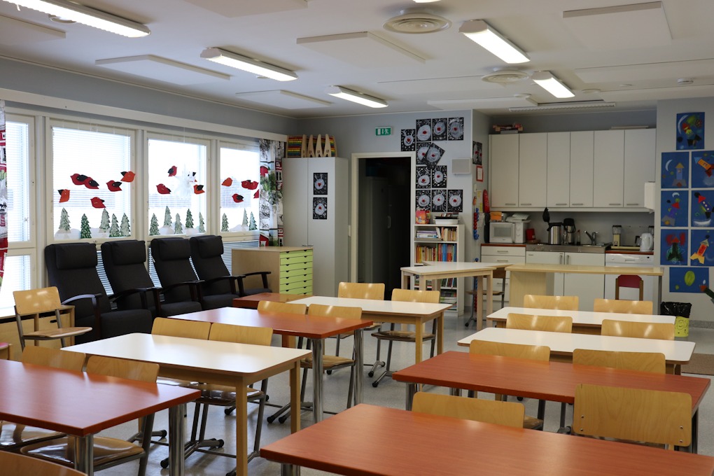 Luokkahuone, jossa taustalla vasemmalla on oviaukko ja vieressä vaalea kaapisto seinällä ylhäällä ja työtaso keskellä. edessä on seitsemän pareittain istuttavia oppilaspöytiä.