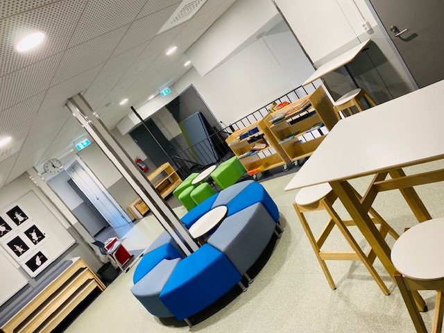 Aseman koulun sisätilojen kalusteita: puisia tuoleja ja pöytä sekä siniharmaita matalia kangasverhoiltuja sohvaistuimia keskellä ympyrän muodossa.