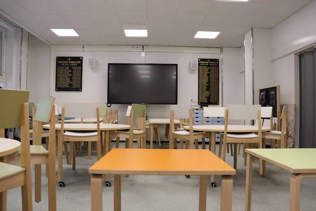 Luokkatila, jossa iso näyttö takaseinällä ja valkoisia tuoleja ja pöytiä ryhminä. Edessä on kaksi suorakaiteenmuotoista pöytää, joista toinen on vaalean oranssi ja toinen vaalean vihreä.
