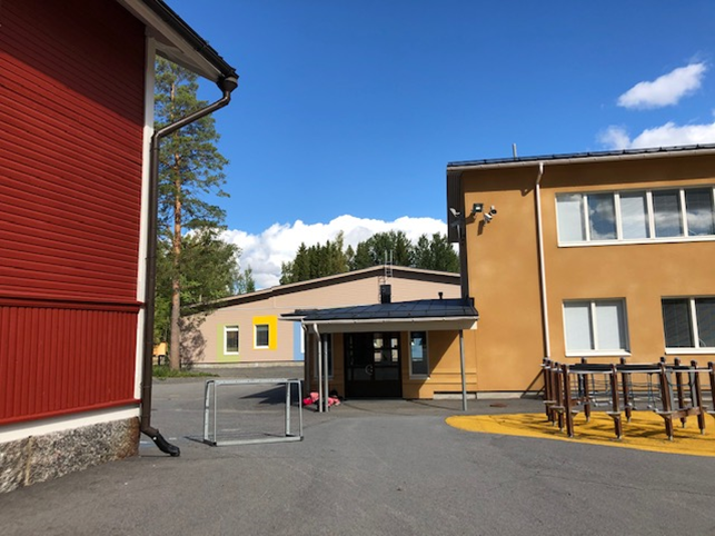 Niemistön koulun pihasta kesällä otettu kuva, jossa edessä on sisäänkäyntiovi katoksen alla ja oikealla matala kiipeilyteline koulun edessä. Vasemmalla näkyy puinen, punainen vanha koulurakennus.