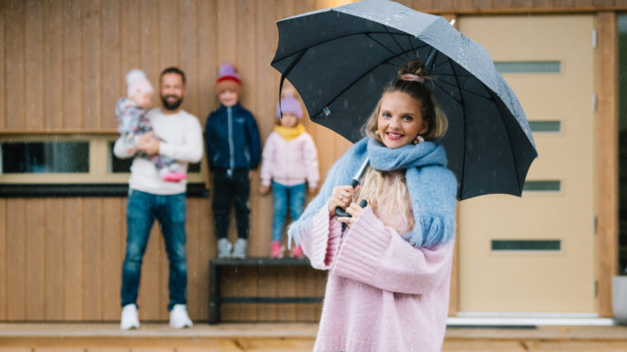 Nainen seisoo sateenvarjon alla ja tuastalla mies ja kolme lasta katoksen alla.