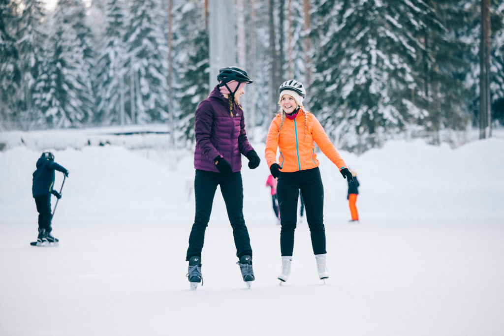 Kuvassa kaksi henkilöä luistelee. Taustalla lapsi kiekkoilee ja muita luistelijoita