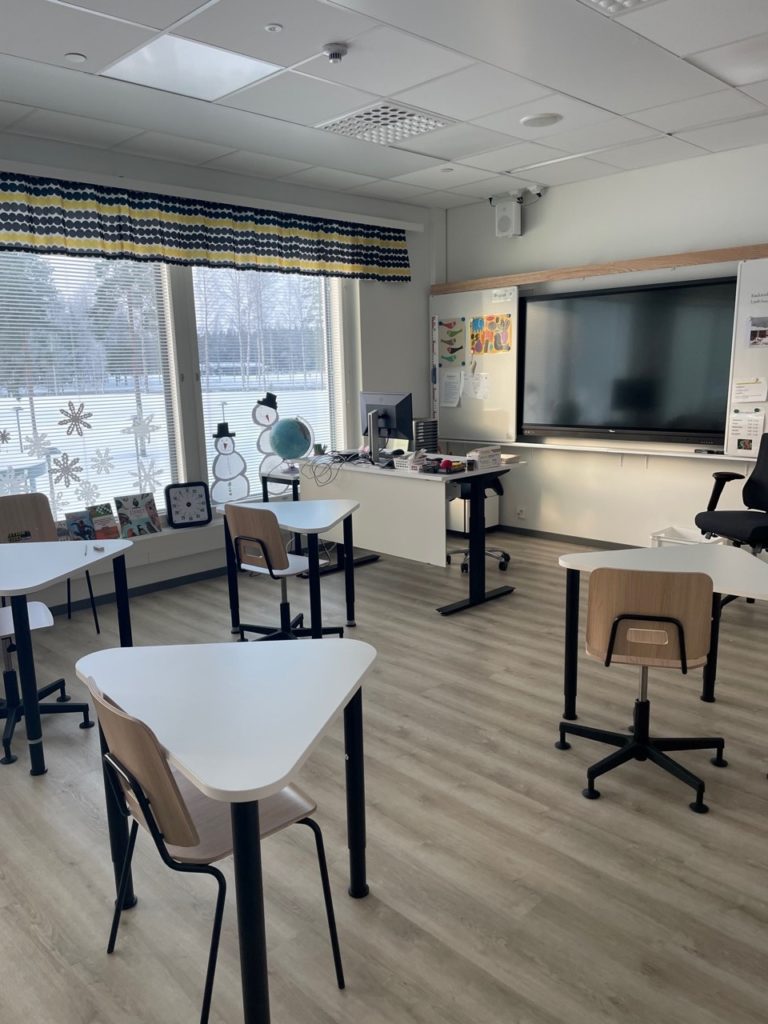 Luokkahuoneessa pulpetteja ja tuoleja, ikkunasta näkyy tavlvinen luminen maisema