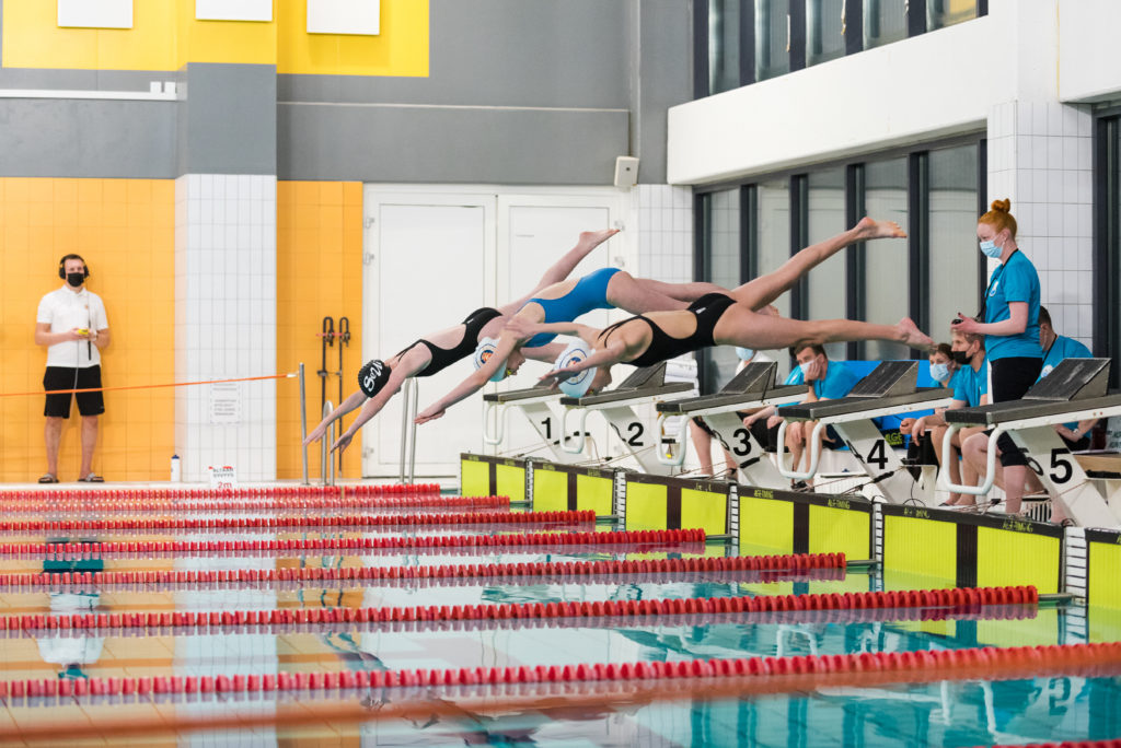 Kuvassa kolme kilpauimaria hyppää uima-altaaseen. Taustalla toimitsijoita.