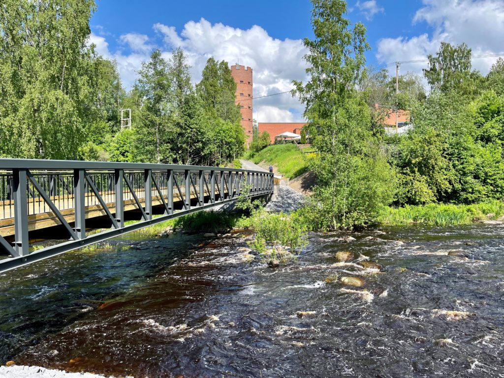 Puukantinen teräsrakenteinen silta yli kosken. Taustalla vesitorni.