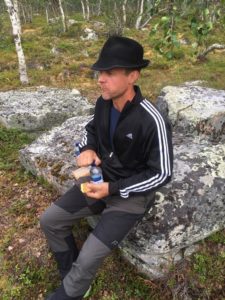 Kuvassa henkilö istuu metsässä kivellä musta hattu päässä ja syö eväitä.