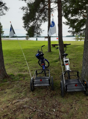 Kuvassa on kaksi sähköpyörää peräkärryineen puistossa järven rannalla. Peräkärryissä on viirit.