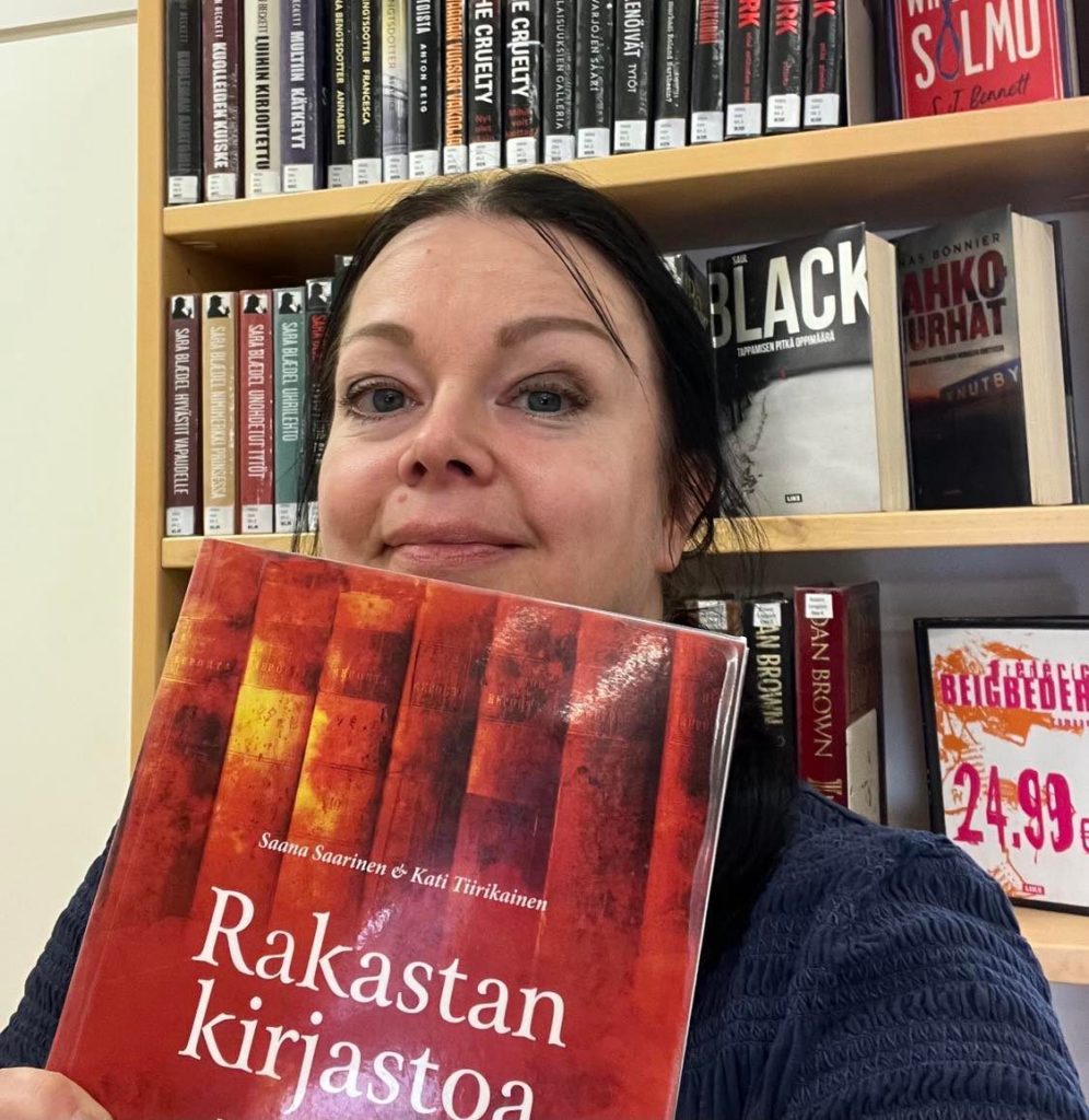 Tekstin kirjoittaja kirjahyllyn edessä, kädessään punasävyinen kirja nimeltään Rakastan kirjastoa.