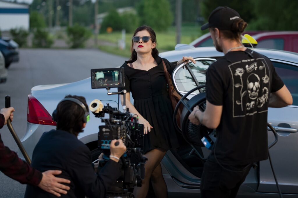 Autosta nouseva aurinkolasipäinen nainen, jota elokuvakamera kuvaa.