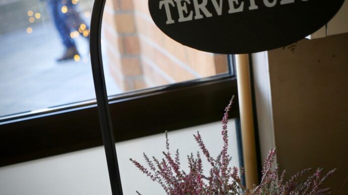 Tervetuloa-kyltin alla kanervia. Taustalla ikkunan takana näkyy kävelijän jalat.