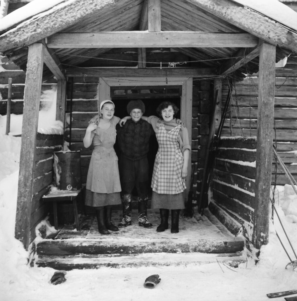 Mustavalkoisessa kuvassa kolme henkilöä vanhan hirsirakennuksen kuistilla talvella.