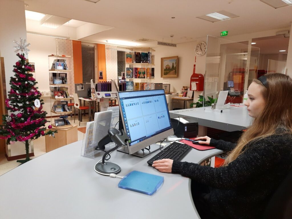 Nuori nainen istuu tietokoneruudun edessä kuvan oikeassa reunassa. Taustalla näkyy kirjastotila.