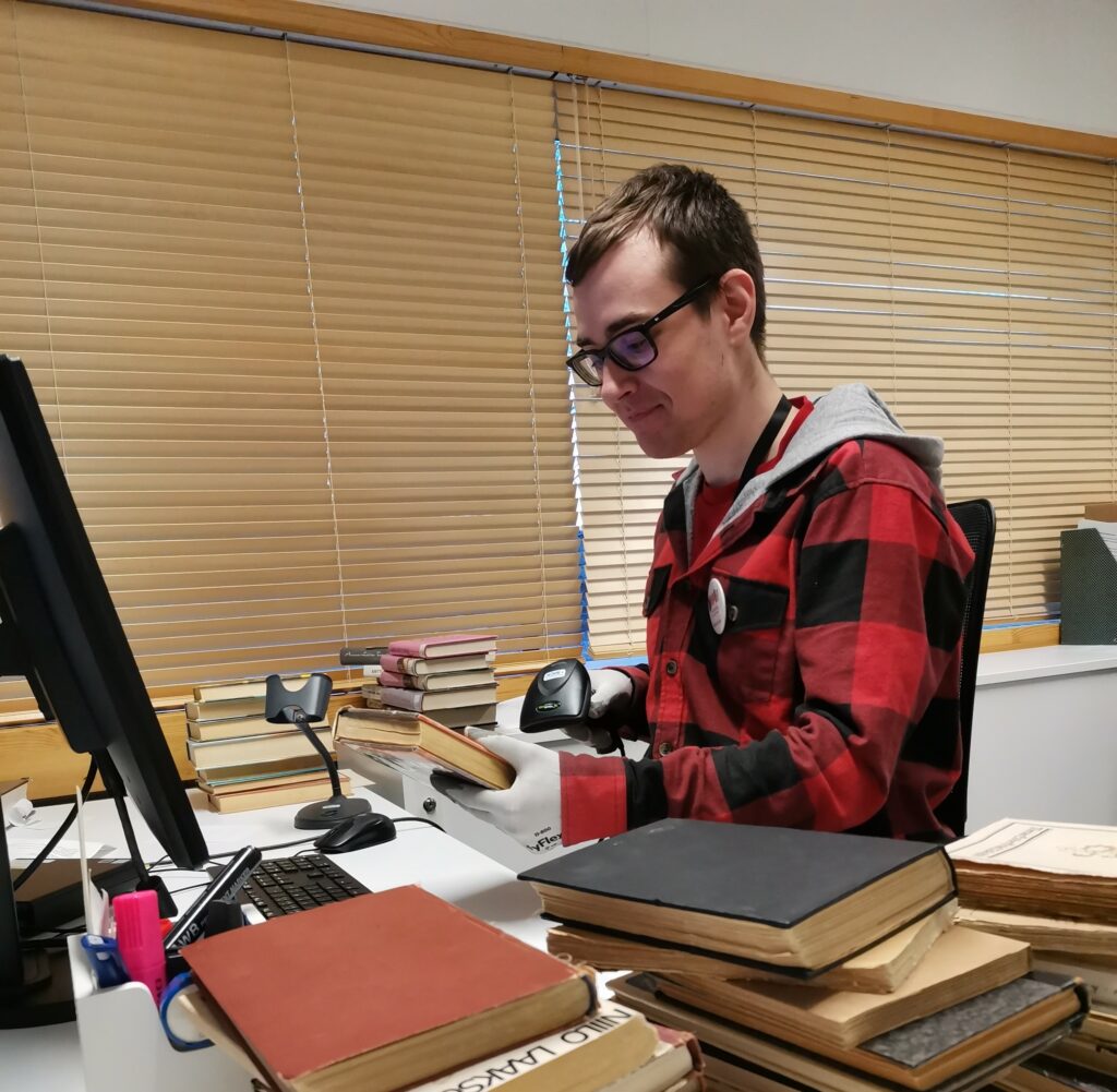 Etualalla kirjakasoja ja niiden takana punamustaan paitaan pukeutunut mies, jolla on kädessään viivakoodin lukija ja kirja.