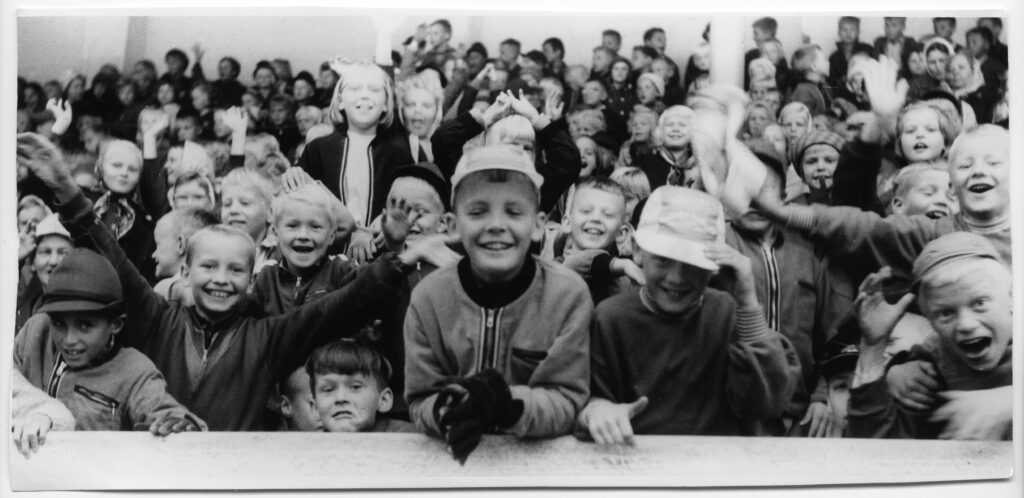 Mustavalkoinen 1960-luvun kuva, jossa joukko hymyileviä, viittovia ja virnuilevia lapsia katsomassa kohti kuvaajaa.