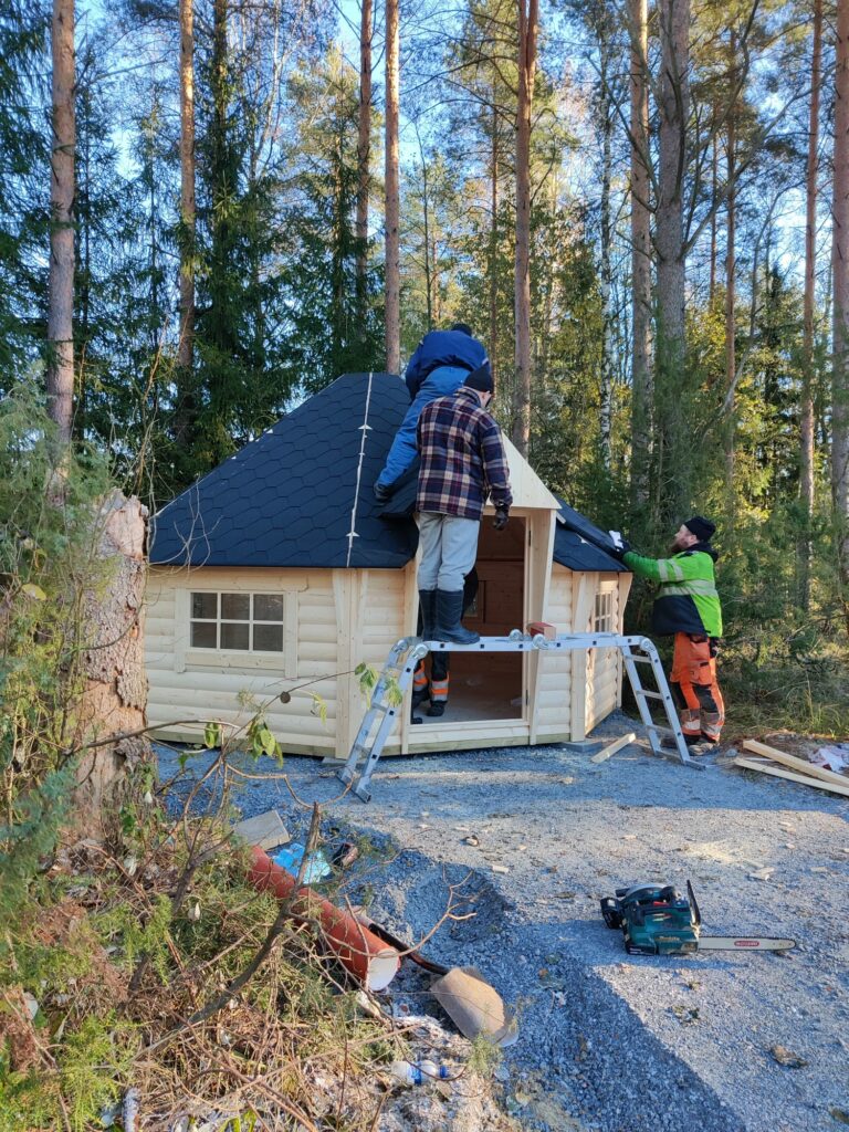 Keskeneräinen kota, jonka katolla kaksi rakentajaa ja oikealla seinustalla yksi.