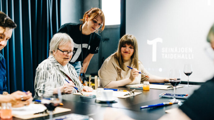 Kolme henkilöä pöydän ääressä maalaustarvikkeiden kanssa