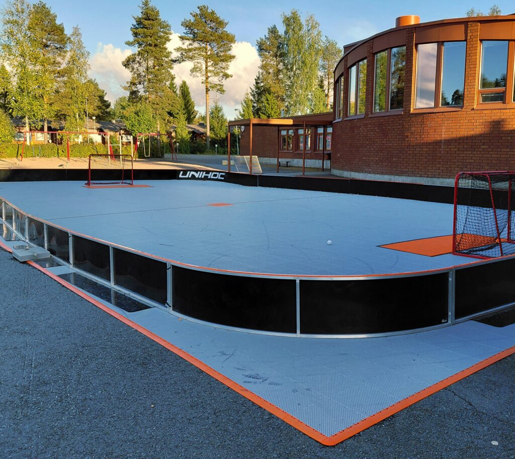 Kuvassa on ulkosalibandykenttä, jossa on vaaleansininen pohja, mustat laidat sekä kaksi maalia, joiden edessä on oranssi alue. Taustalla on puita, kiikkuja sekä Hyllykallion koulurakennus.