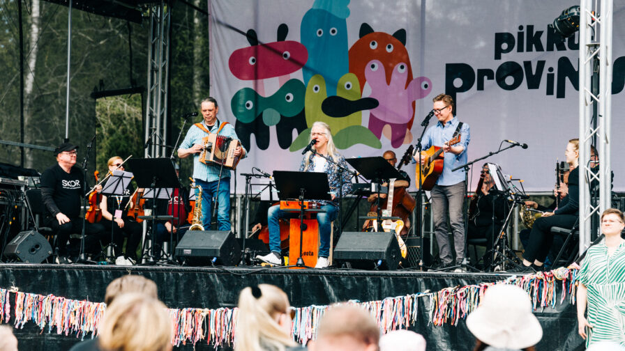 Kuvassa orkesteri esiintyy esiintymislavalla, jonka edustalla on joukko ihimisä.