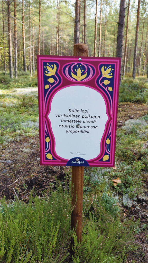 Kuva metsästä, taustalla on puita, kiviä ja varpuja. Keskellä kuvaa värikäs opastetaulu, jossa kehotetaan kulkemaan polkujen läpi ja ihmetellen pieniä olentoja luonnossa.