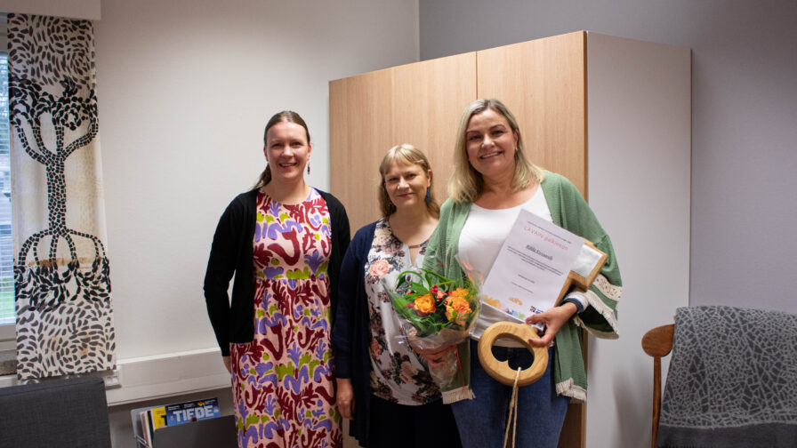 Kuvassa kolme henkilöä seisomassa rivissä. Yhdellä heistä on kädessään kukkakimppu sekä kunniakirja.