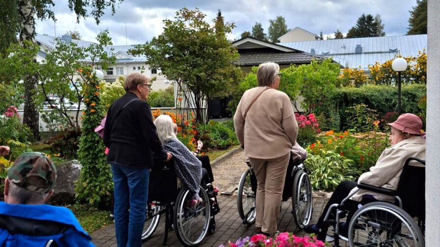 Pyörätuoleissa istuvia heitä ulkoiluttavia ikäihmisiä katselemassa syksyistä puutarhaa.