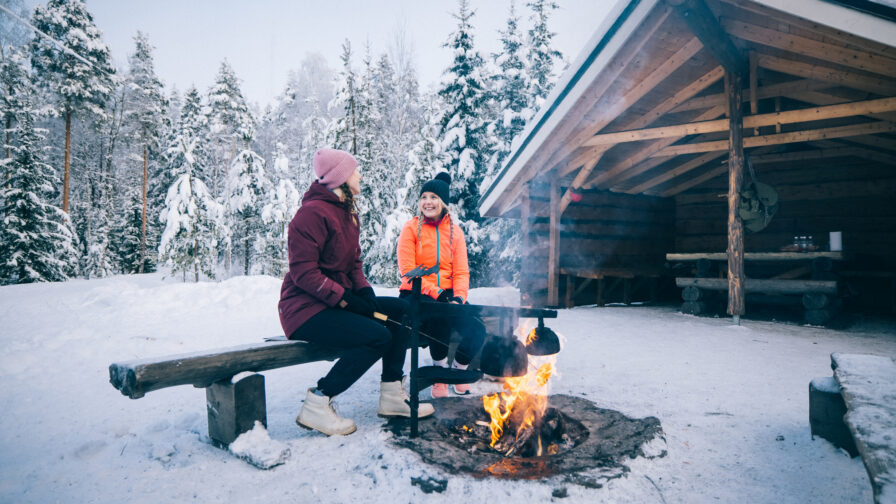 Kaksi henkilöä istuu penkillä nuotiolla. Taustalla lumista metsää ja laavu.