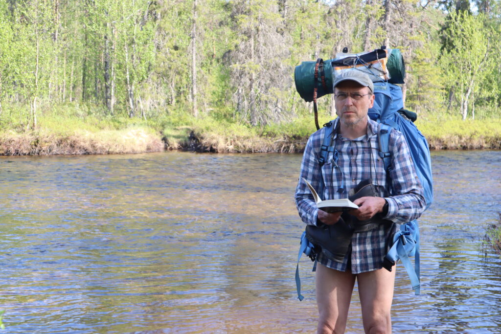 Mies, jolla on rinkka selässä, seisoo joessa kirja kädessään.