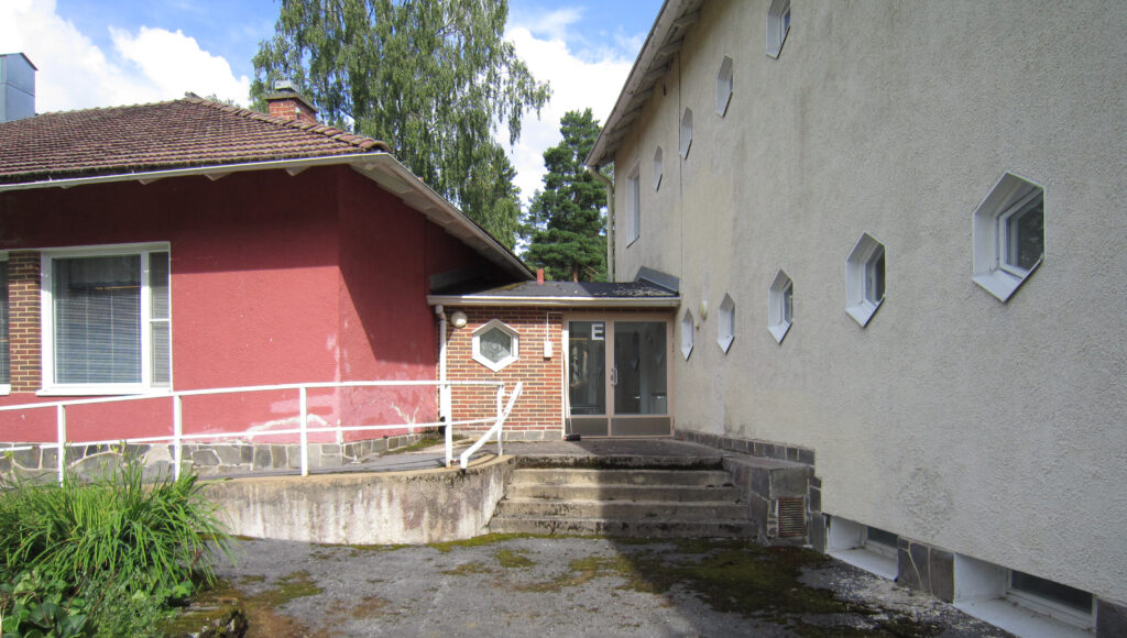 Punaisen ja vaalean rakennuksen välissä on E-ovi. Oven edustalla portaat ja luiska. Vaalean kaksikerroksisen rakennuksen ikkunat ovat pienet ja kuusikulmaiset.