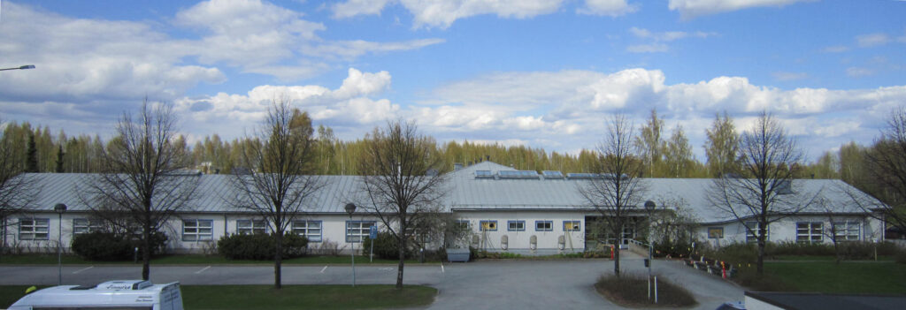 Julkisivukuva Myllyrannan palvelukeskuksesta. Yksikerroksinen rakennus, jossa on vaaleat seinät ja siniharmaa katto, sekä ikkunapuitteet. 