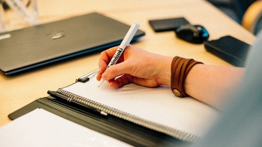 Lähikuva, jossa kädessä on Seinäjoki-kynä, jolla kirjoitetaan muistioon. Taustalla pöydällä on suljettu tietokone ja langaton hiiri.