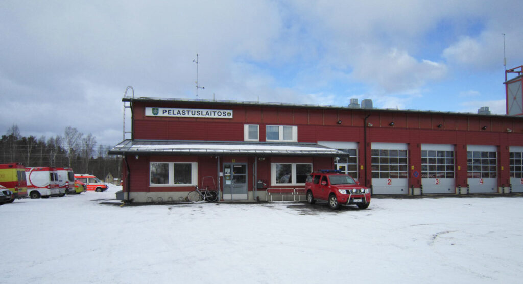 Nurmon pelastuslaitoksen rakennus, jonka pihassa on paloaseman johtoauto. Punainen rakennus, jossa näkyy viisi valkoista nosto-ovea. 