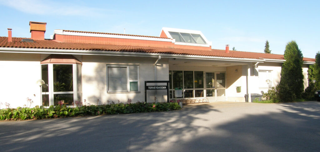 Terveysasema on matala rakennus, jossa valkoiset seinät ja punainen katto. 